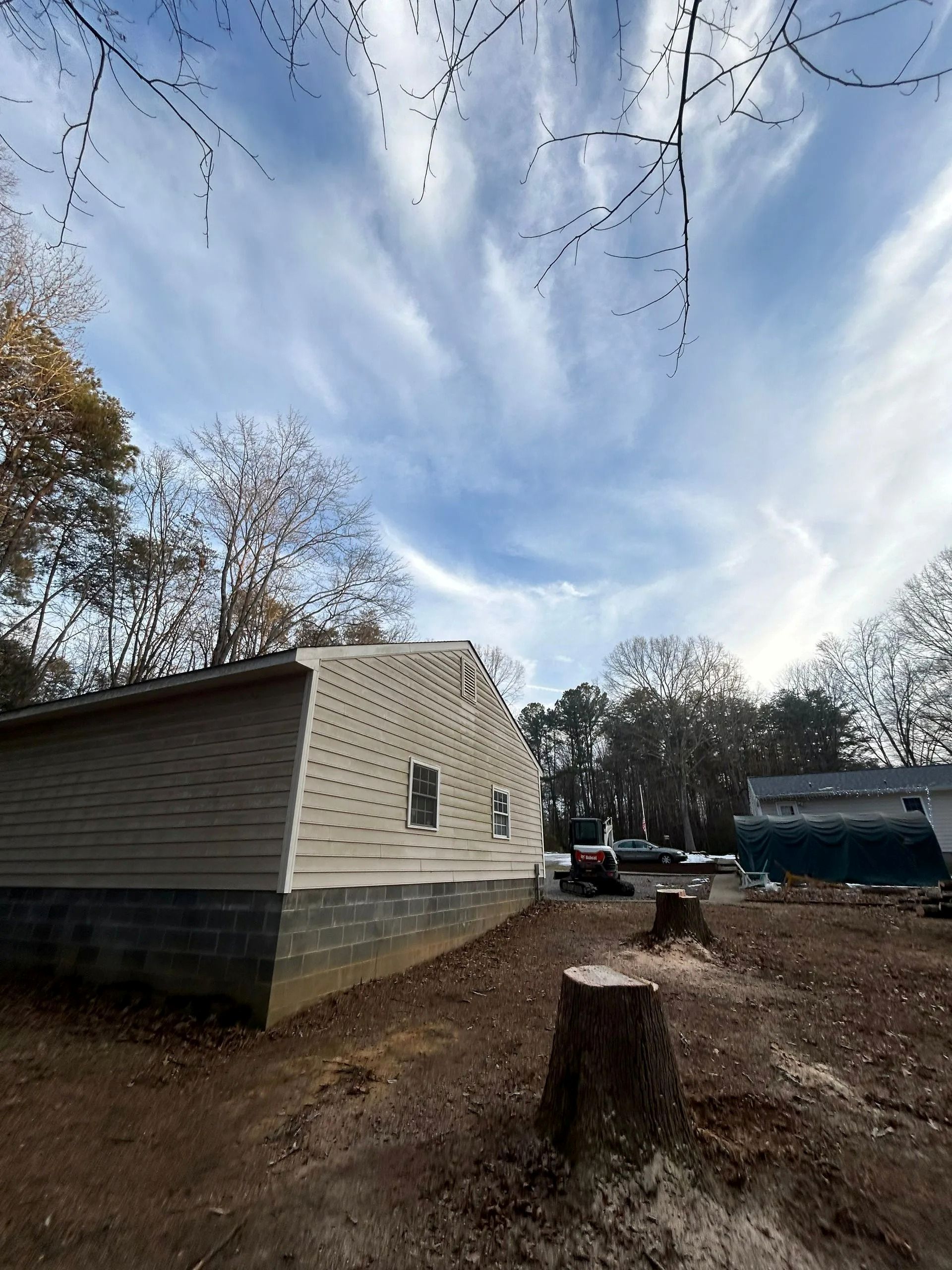 A tan, side-gabled house with a grey concrete block foundation sits in a yard with a tree stump under a bright blue sky.