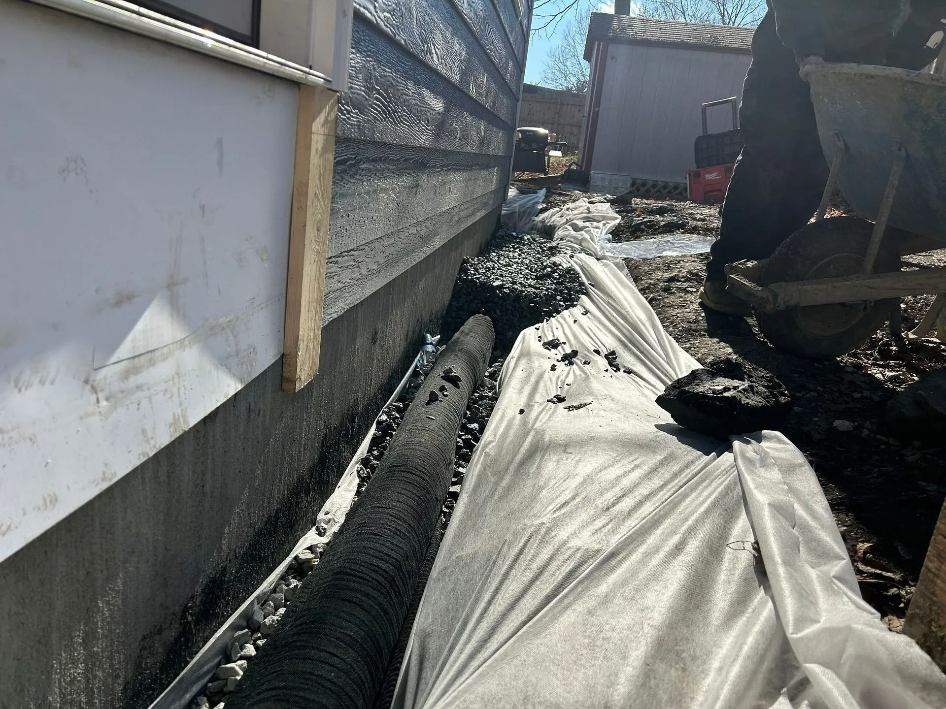 A person uses a wheelbarrow next to a foundation wall where a perforated drainage pipe sits on top of landscaping fabric.