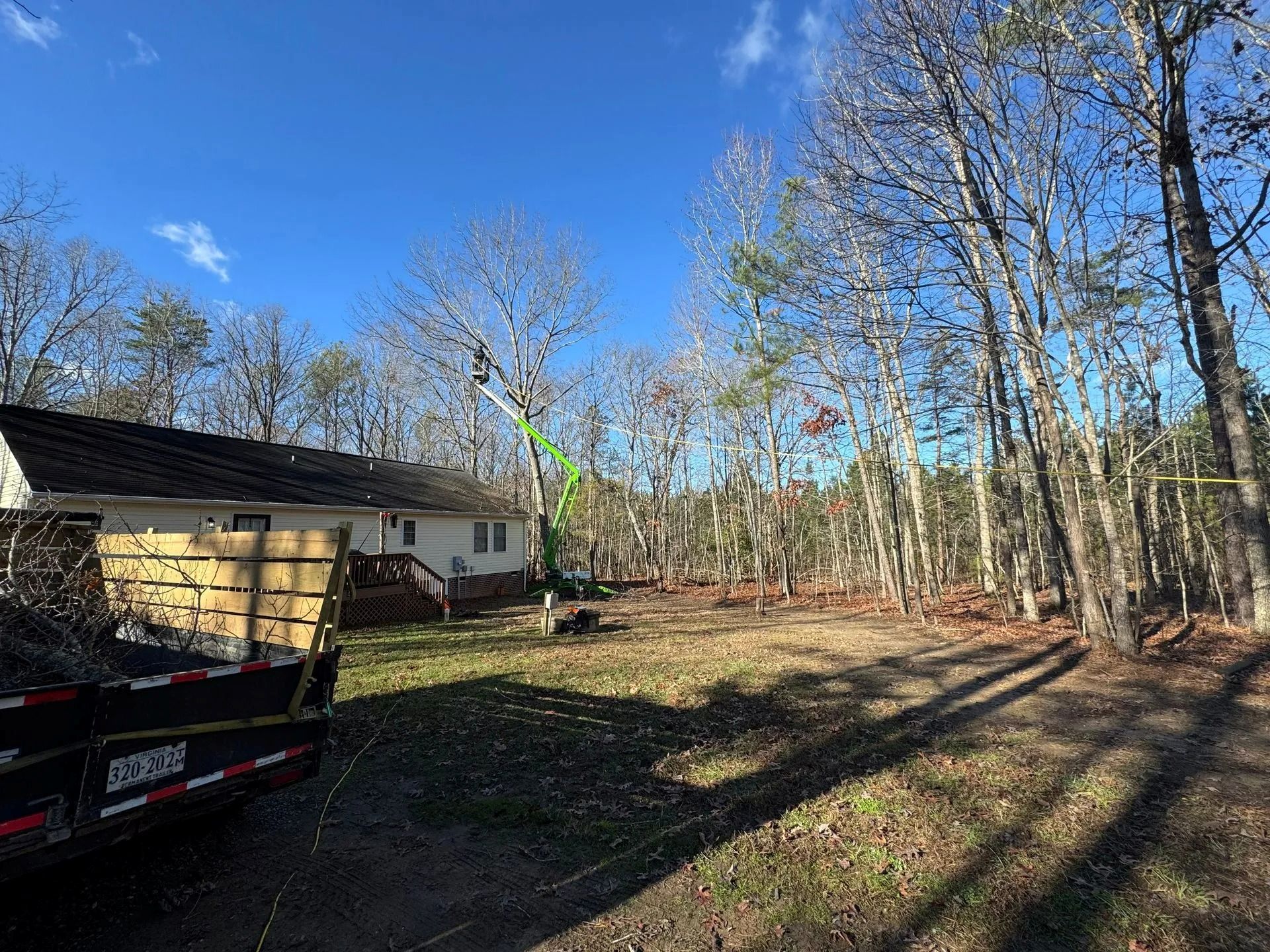 A worker in a bucket truck prunes trees near a house, with a debris trailer parked in the foreground.