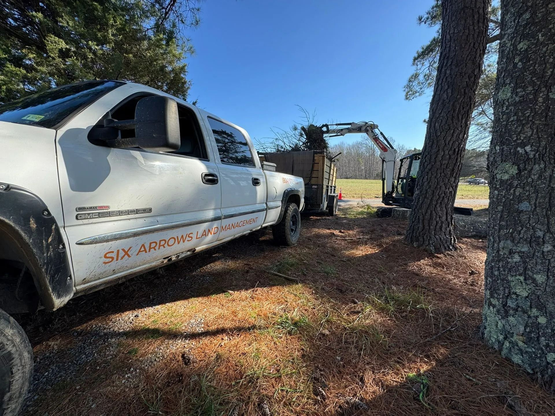 A white pickup truck parked by a tree, with an excavator loading debris into a trailer in the background on a sunny day.