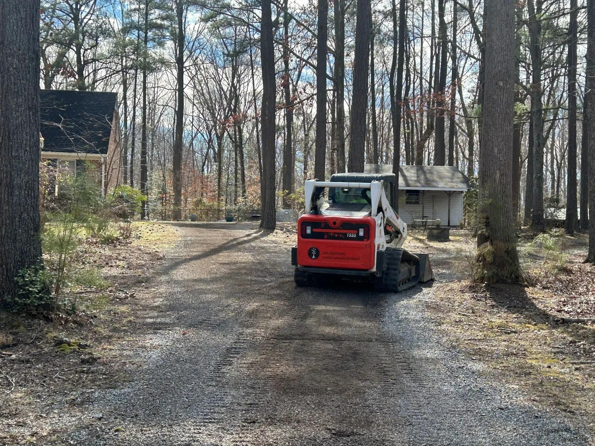 A red and white Bobcat skid steer loader sits on a gravel driveway in a wooded area near two small residential buildings.