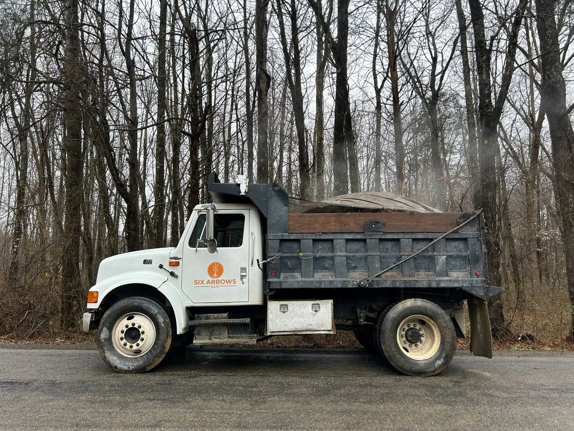 A white dump truck with an orange logo is parked on an asphalt road in front of a dense forest.