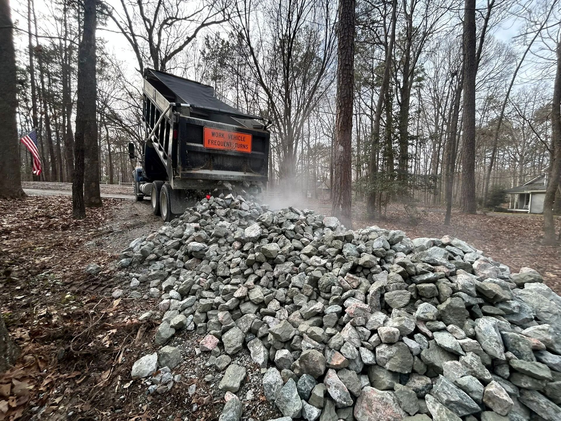 A dump truck emptying a large pile of gray, jagged rocks onto a dirt path in a wooded area.