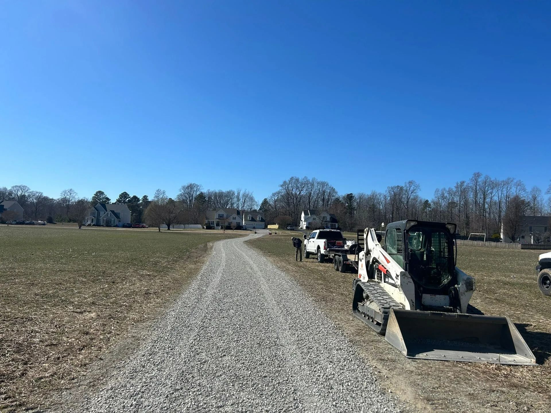 A gravel path leads through an open field toward houses, with a white pickup truck and a white skid-steer loader nearby.