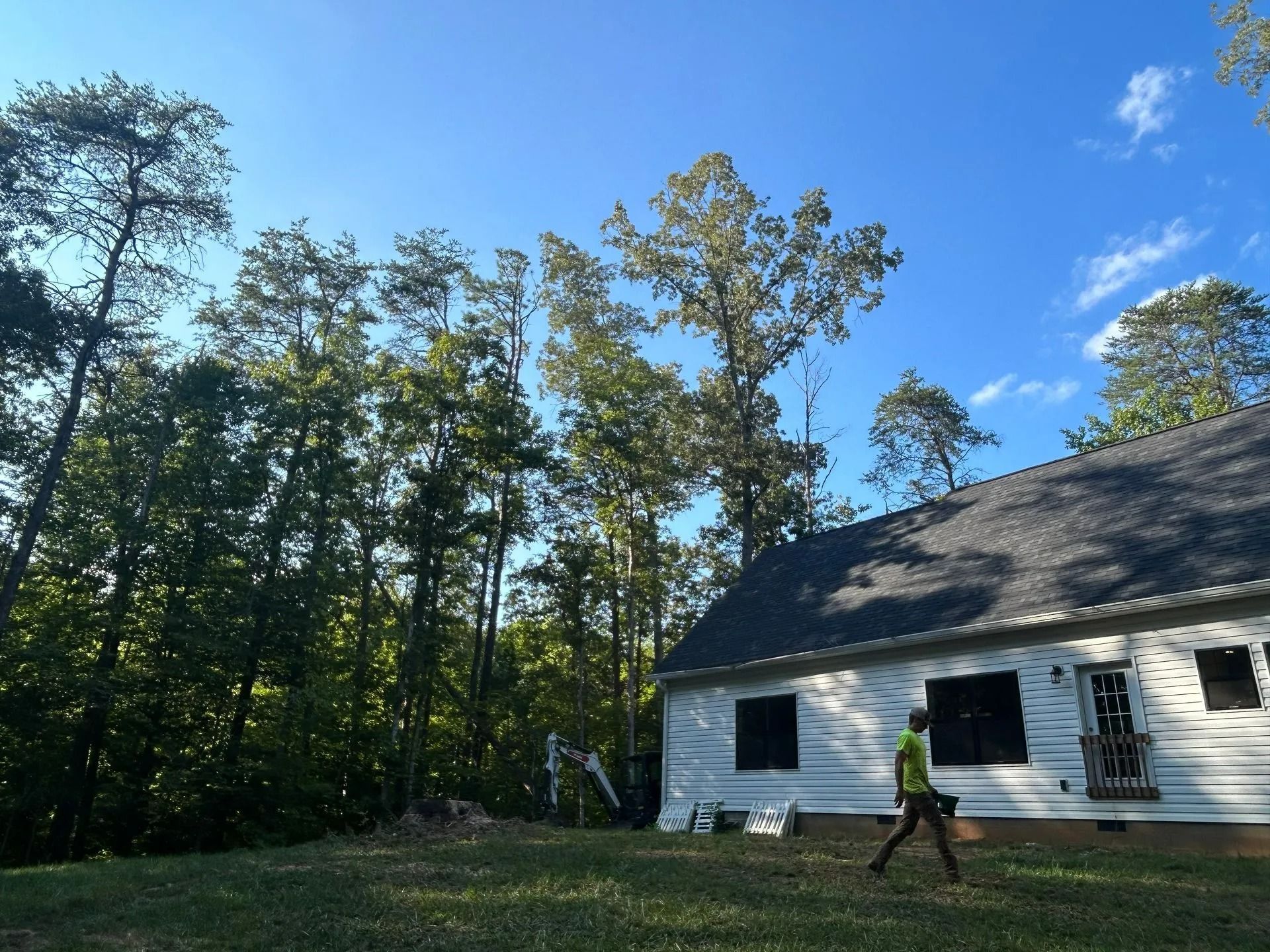 A person in a bright yellow shirt walks across a grassy lawn past a white house near a dense forest under a blue sky.