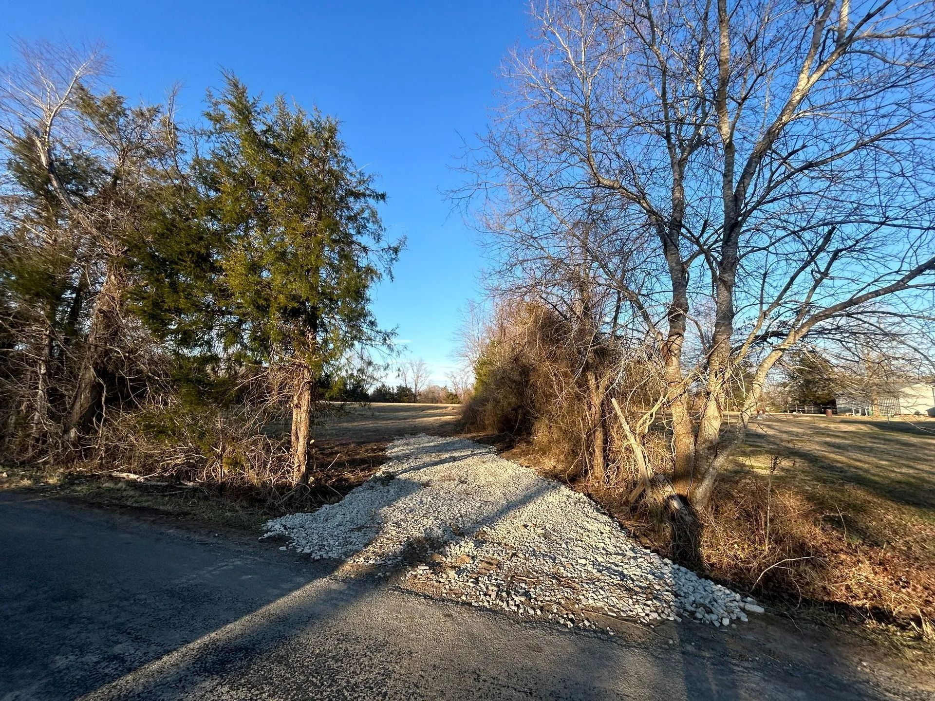 A gravel driveway entrance leads from an asphalt road into an open field framed by trees under a clear blue sky.