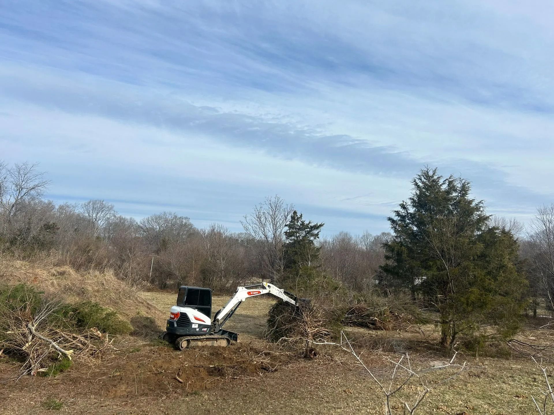 A small white Bobcat excavator clears brush and piles debris in an open field under a blue, cloudy sky.