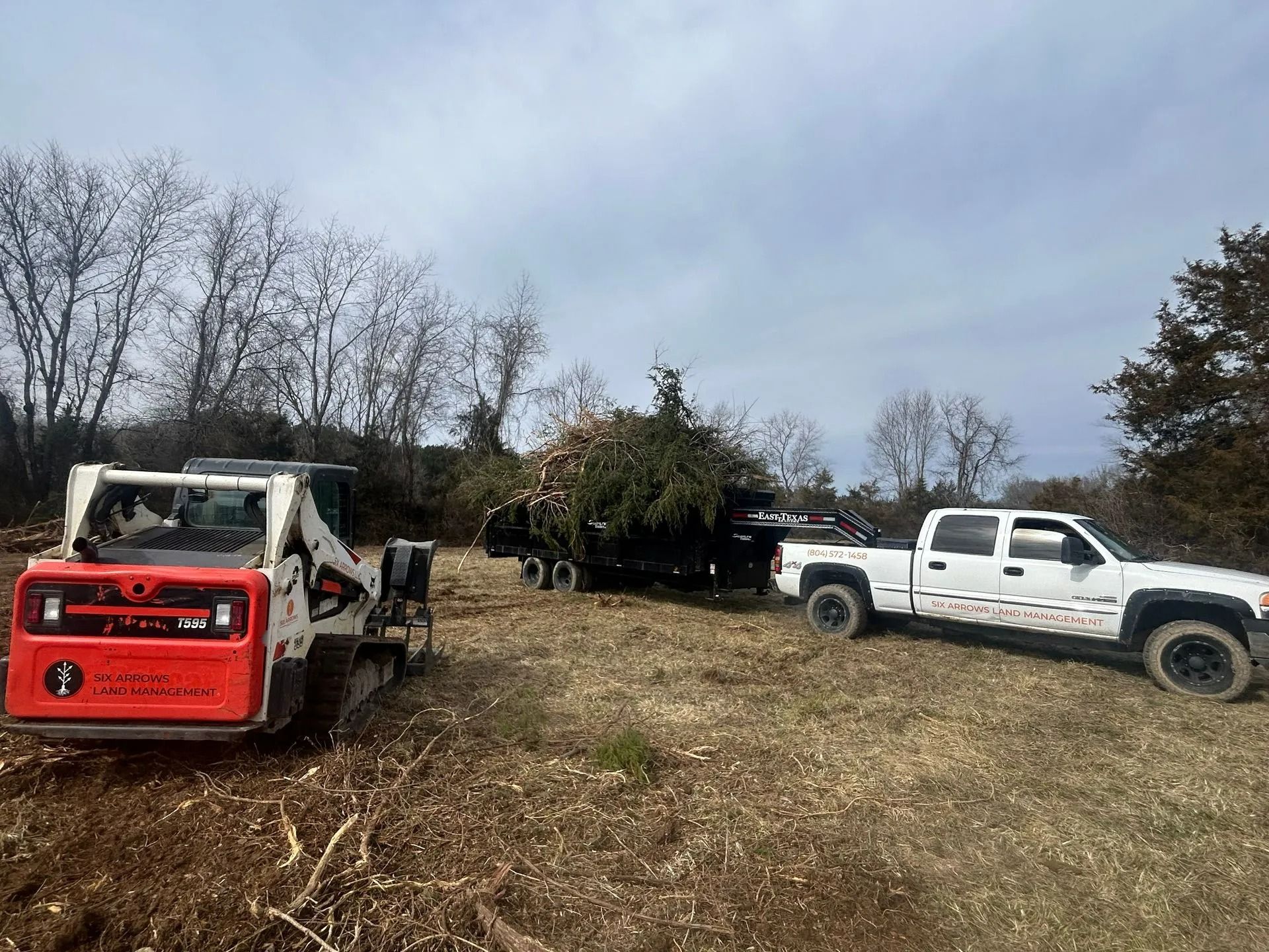 A red skid steer, a white pickup truck, and a trailer loaded with brush in a cleared, grassy field under a cloudy sky.