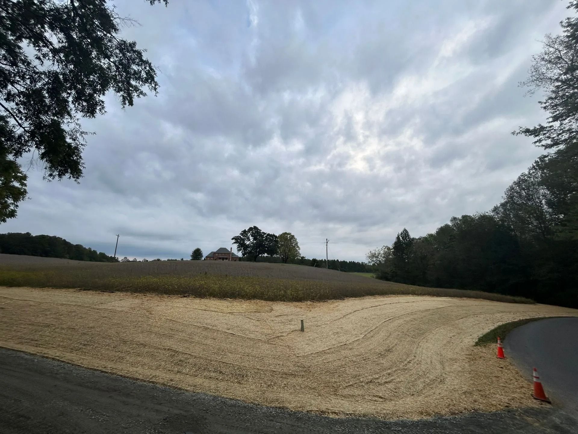 A dirt road leads past a patch of fresh straw mulch on a grassy hillside under a cloudy sky.