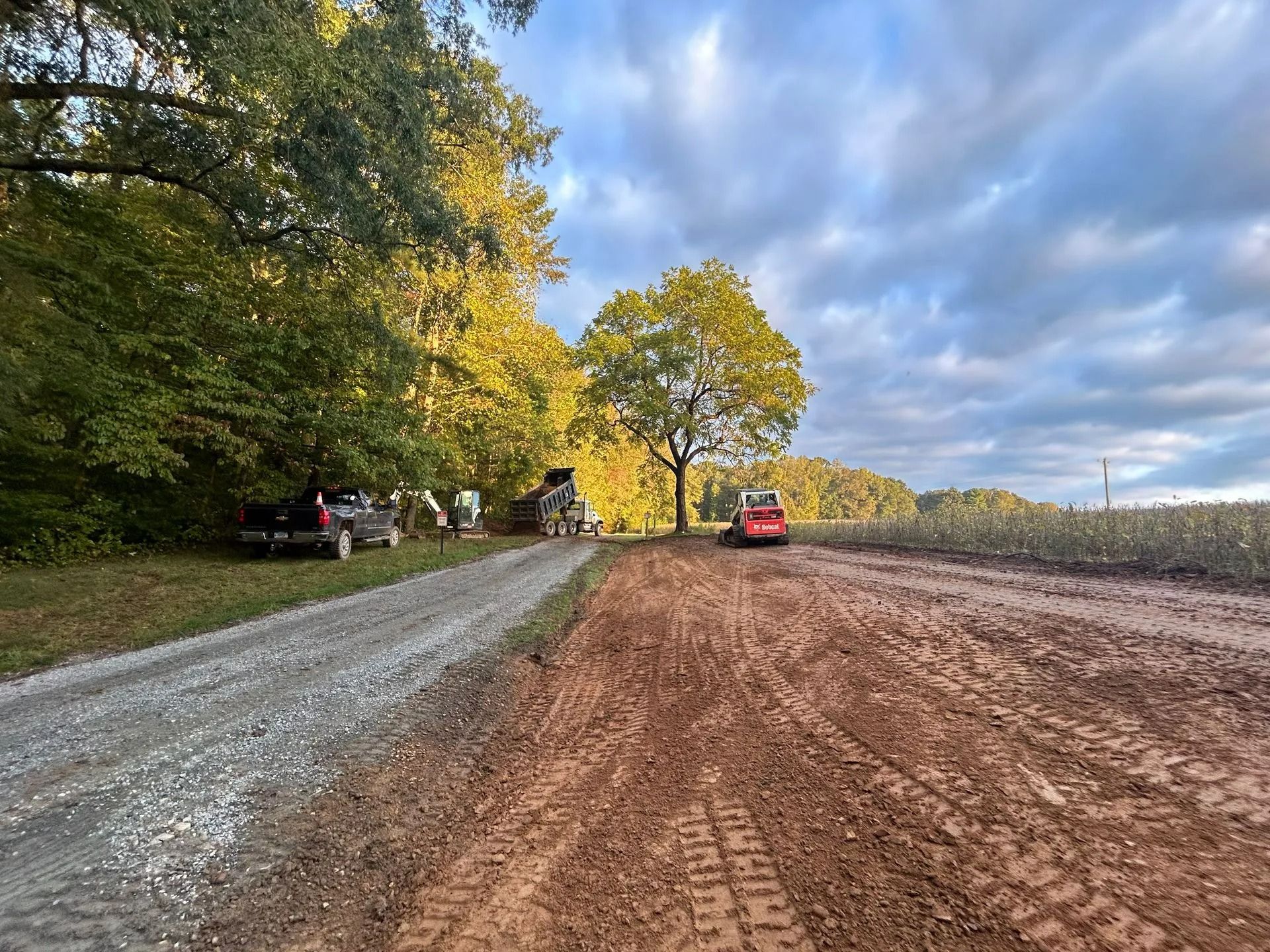 A gravel road leads past a wooded area and a dirt field, with a pickup truck and a small construction vehicle parked nearby.