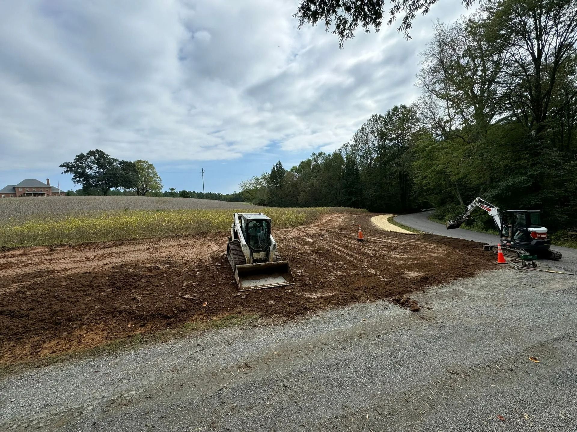 Construction equipment clears dirt and soil on a site near a gravel driveway and tree line under a cloudy sky.