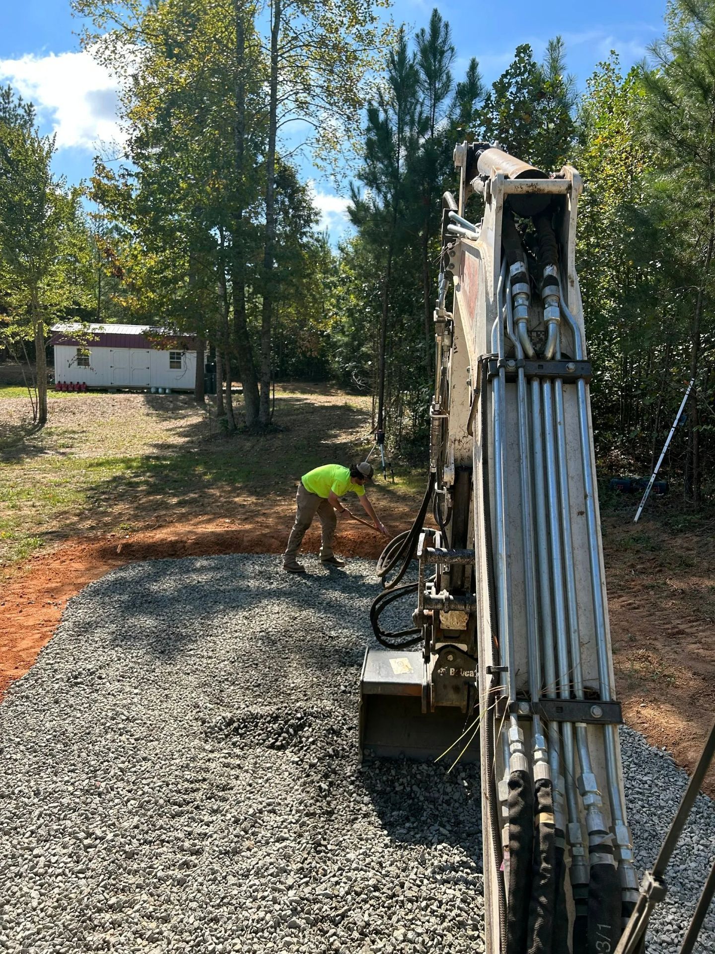 A construction worker in a lime green vest stands near a heavy excavator on a gravel-covered site by a wooded area.