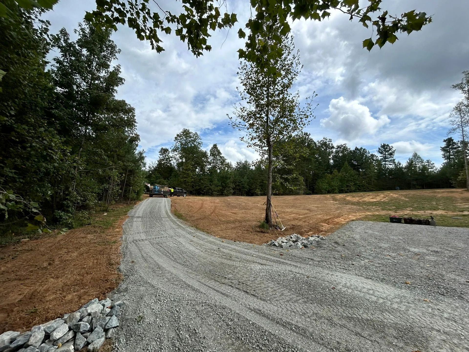A gravel driveway curves through a cleared, grassy lot toward a wooded area under a blue sky with white clouds.