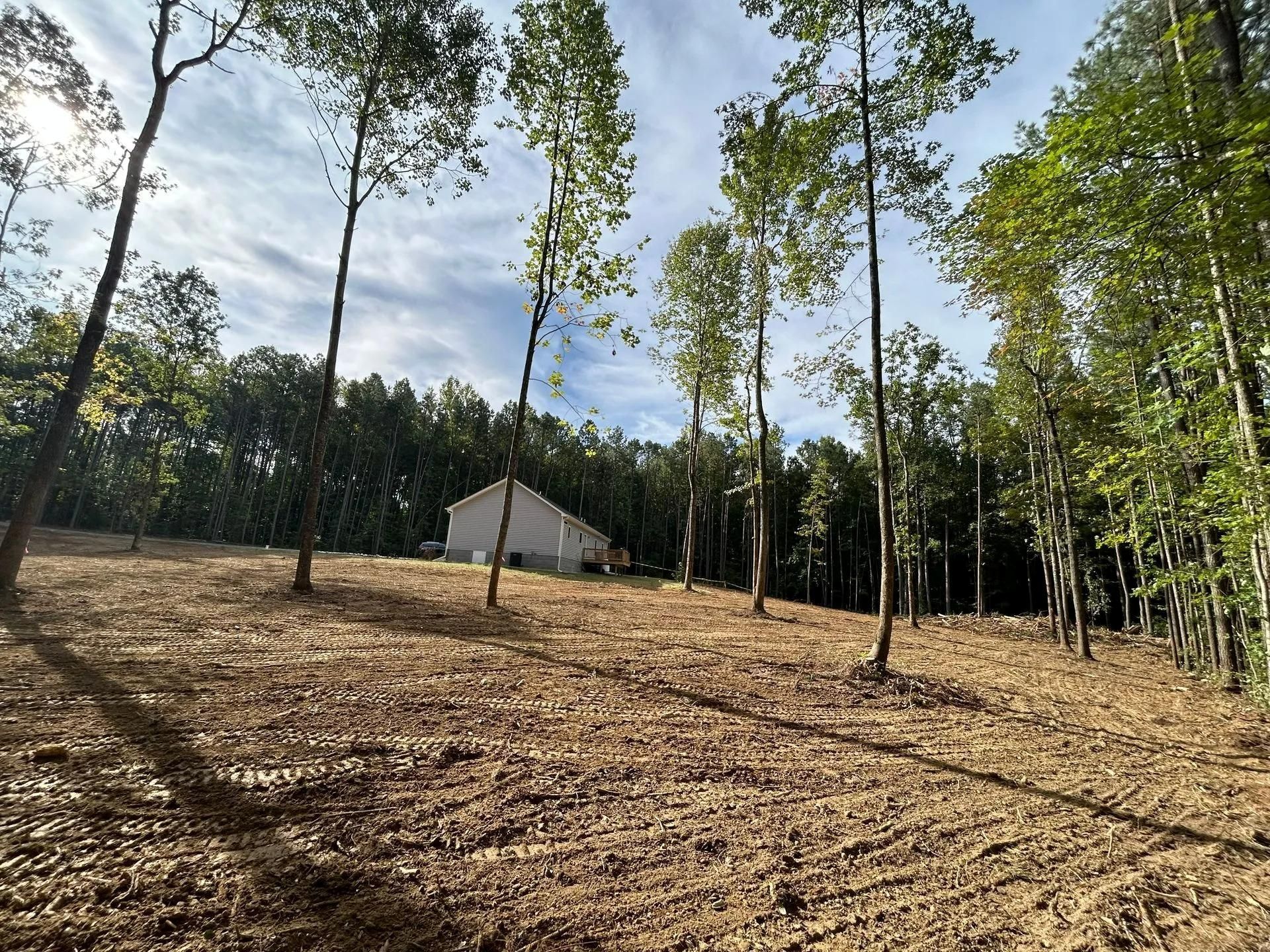 A partially cleared, hilly wooded lot with a single-story house visible in the distance under a sunny, partly cloudy sky.