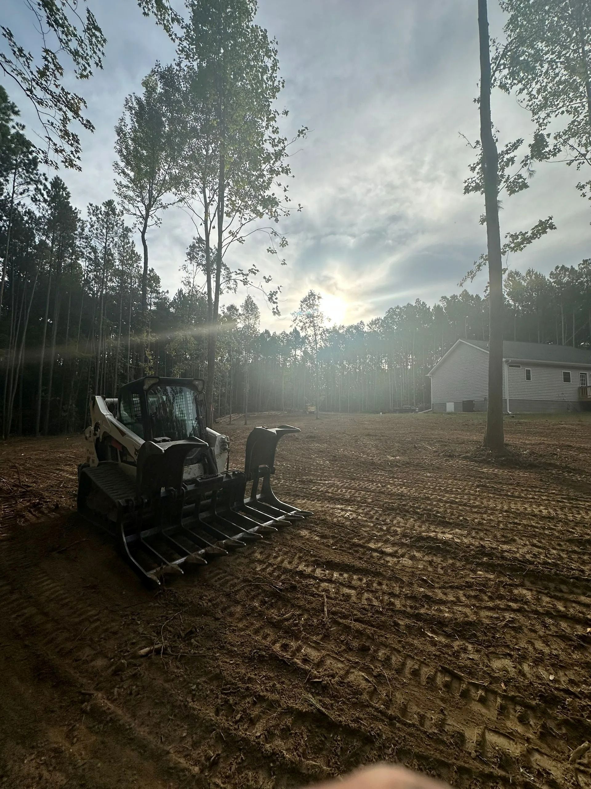 A white skid steer with a rake attachment parked in a cleared dirt lot at sunset, near a house and trees.