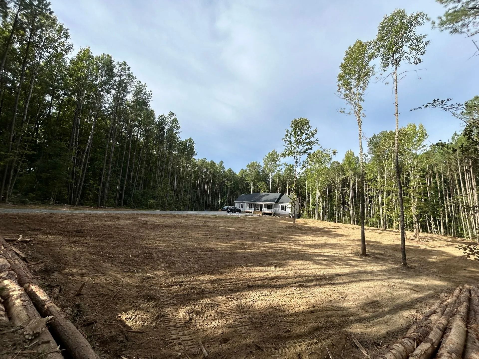 A clearing of dirt and mulch in a wooded area, with a house visible in the distance under a bright sky.