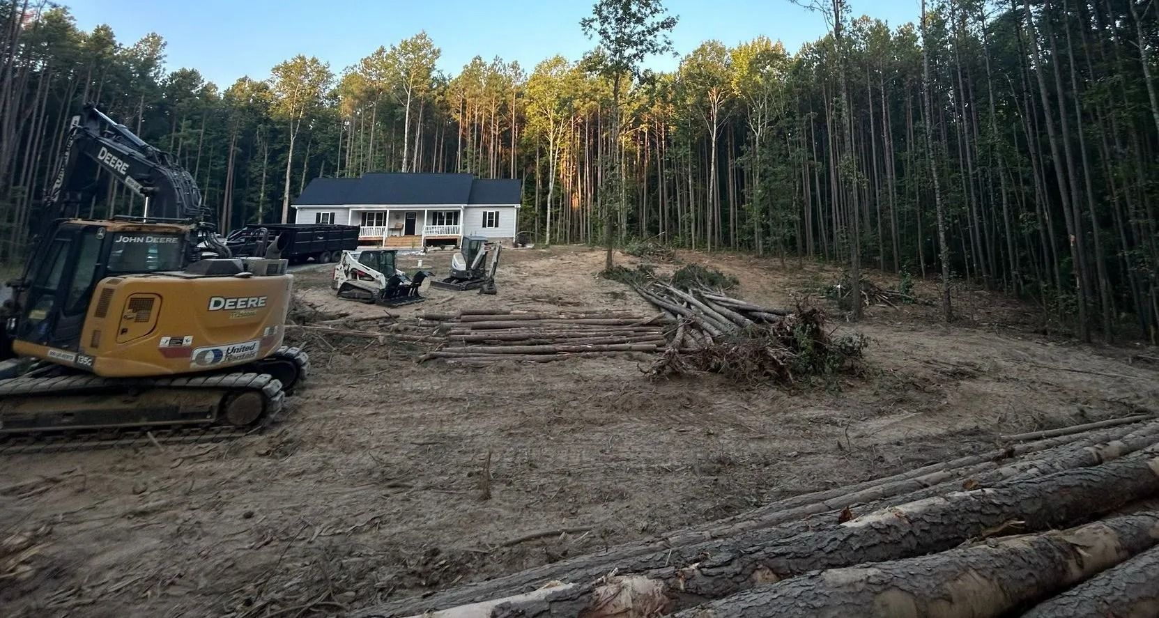 A construction site with heavy machinery, piles of logs, and a white house in the background amid a forest.