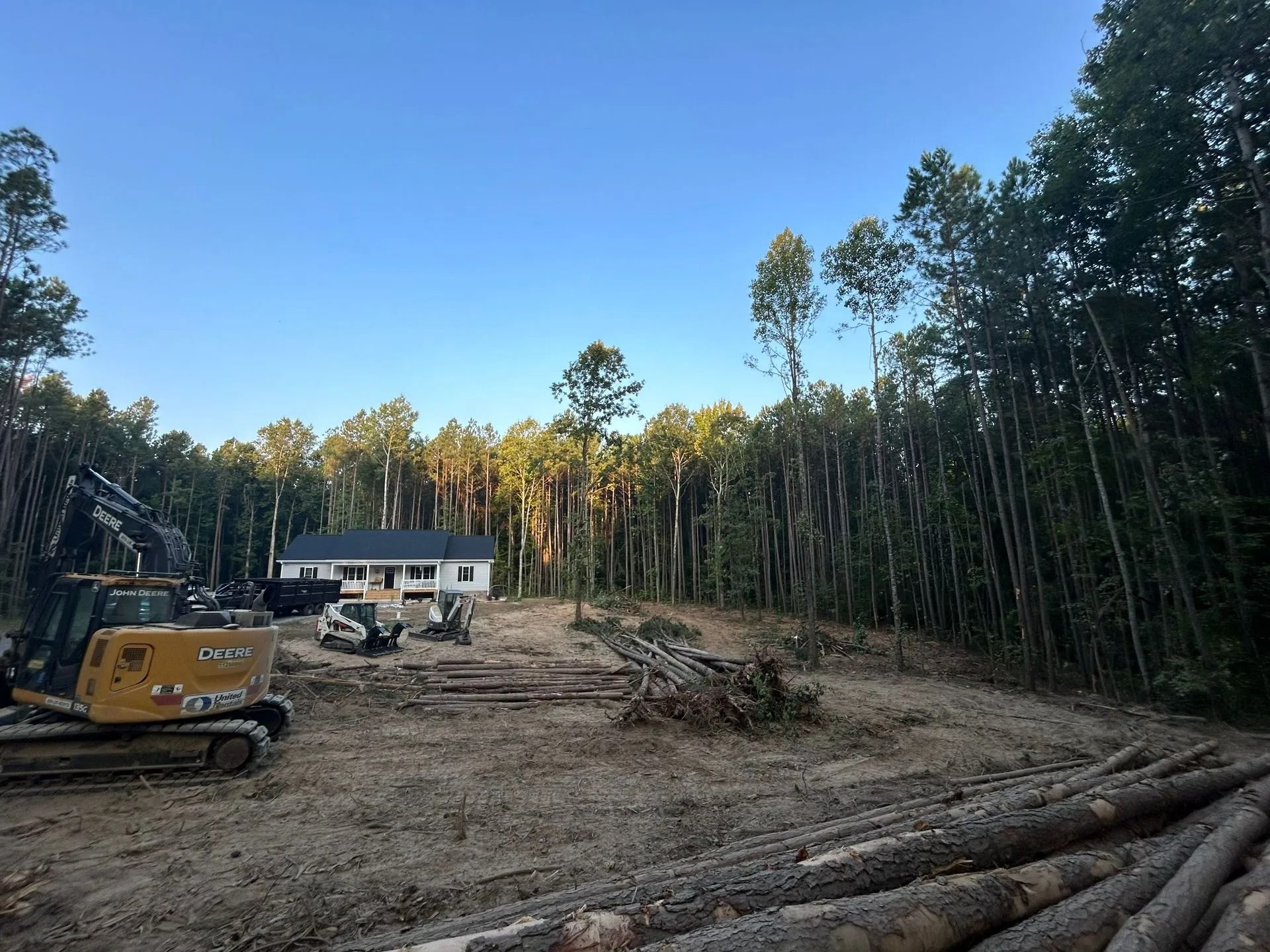 Construction equipment sits on a cleared, dirt-covered construction site in front of a white house surrounded by trees.