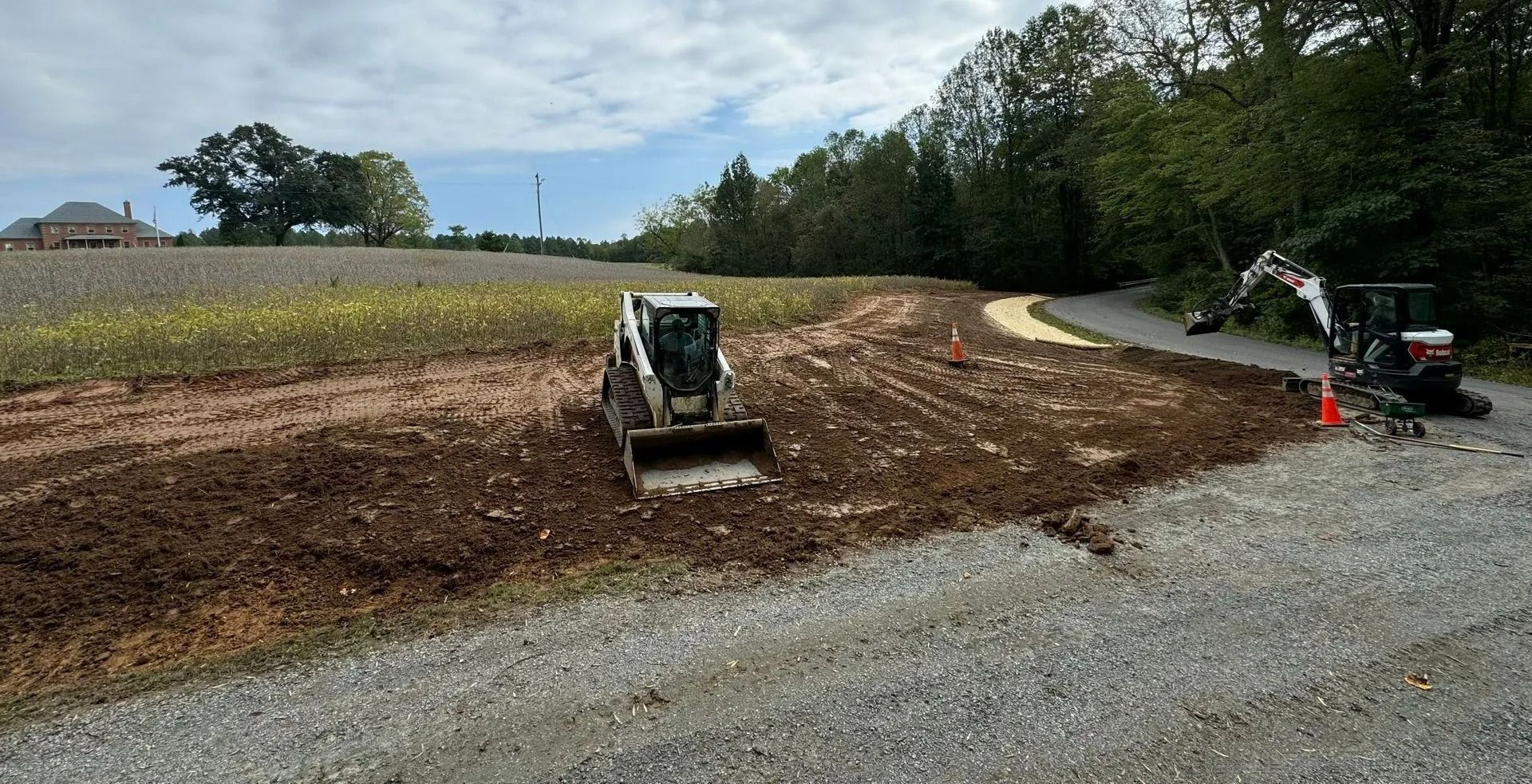 A skid steer and an excavator work on a dirt lot near a gravel road, with a field and trees in the background.