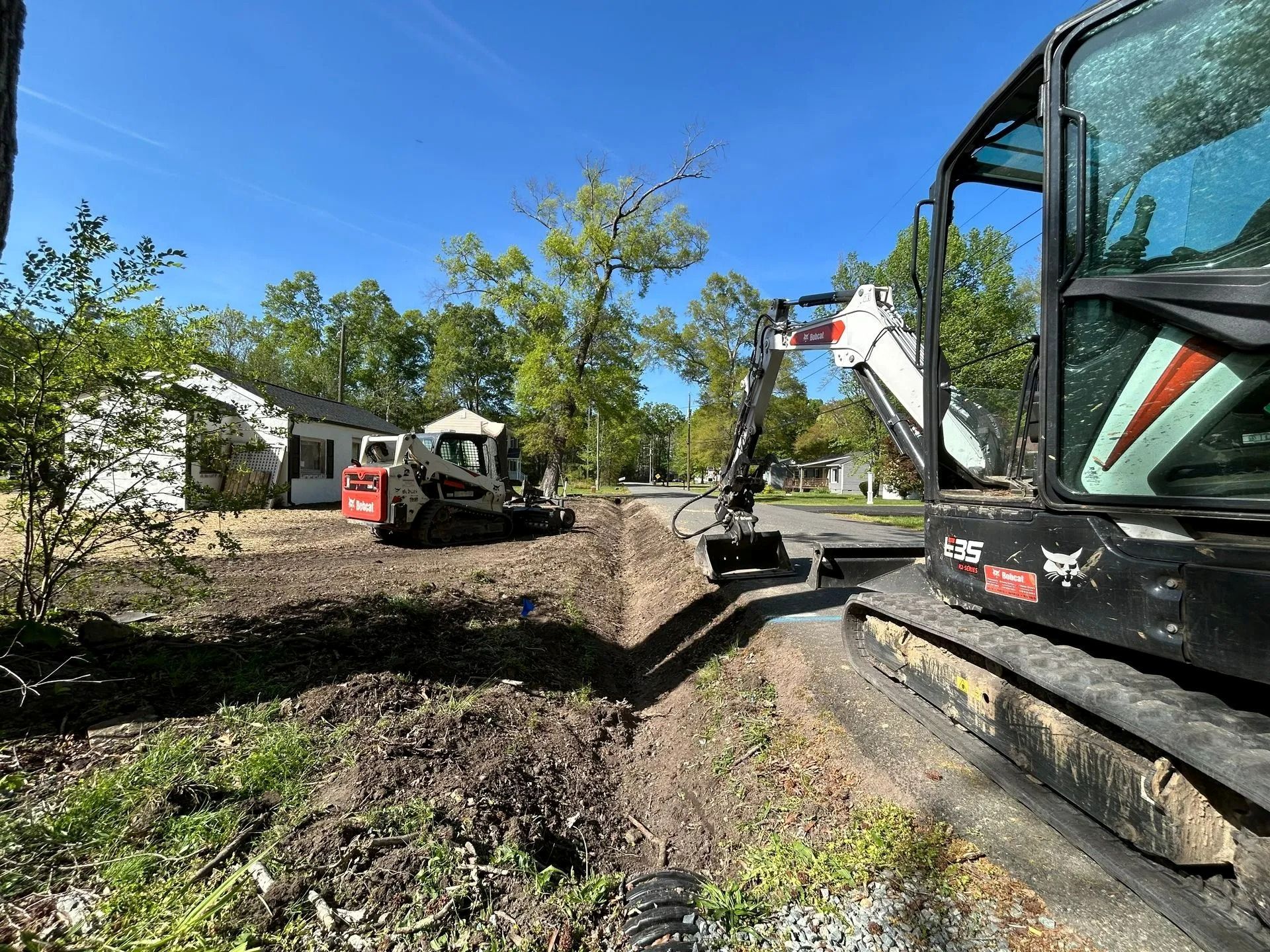 Two Bobcat construction machines dig a trench along a residential street under a clear blue sky.