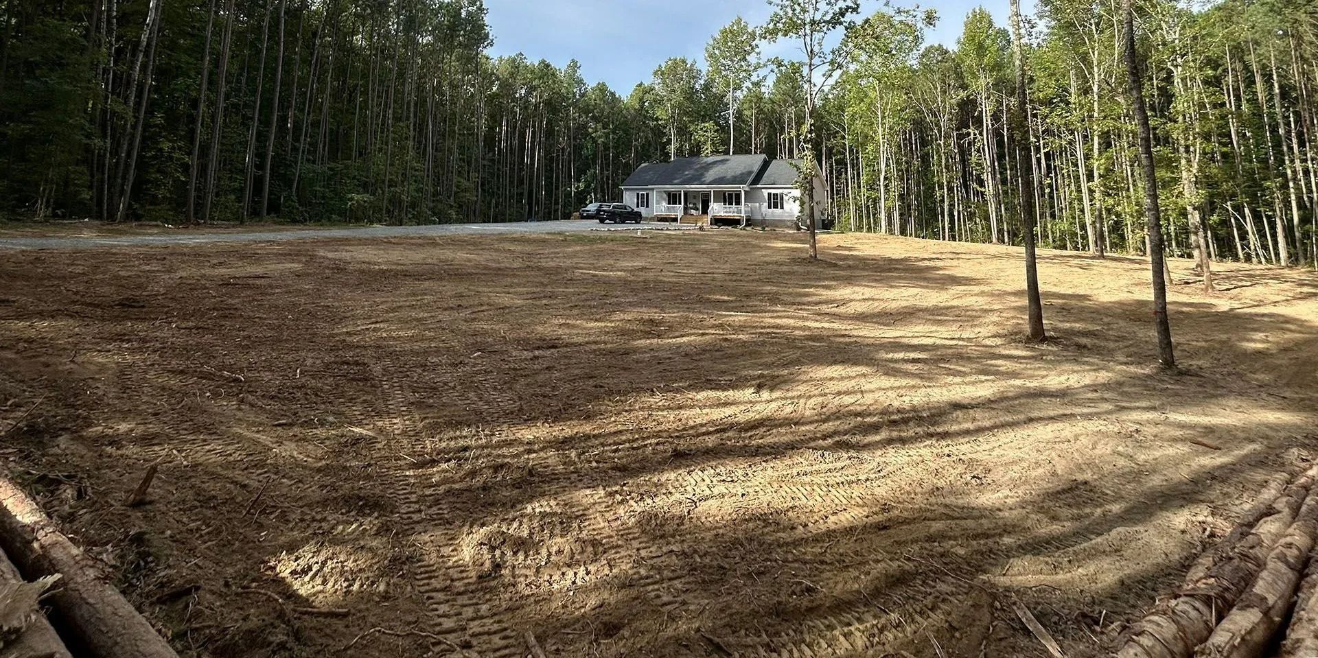 A house sits in the distance behind a large, cleared plot of dirt surrounded by a dense forest of trees.