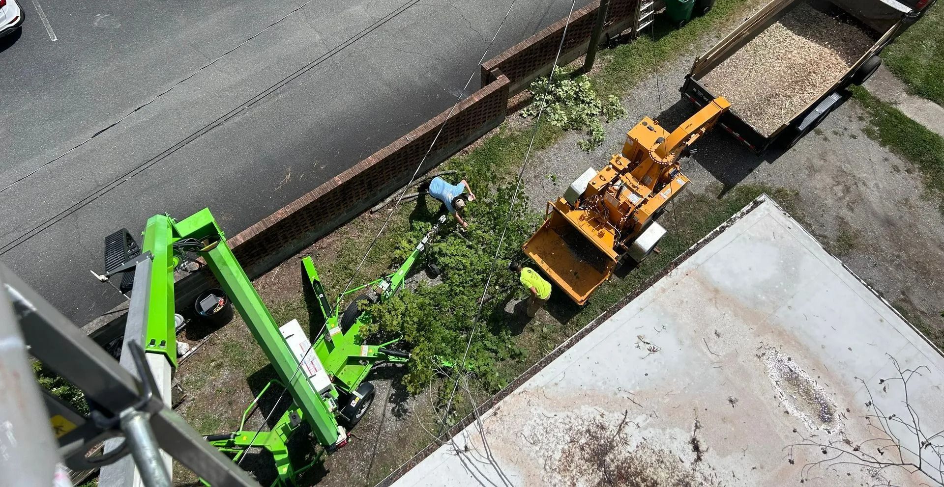 High-angle view of a bright green cherry picker and an orange wood chipper working next to a trailer on a grassy verge.