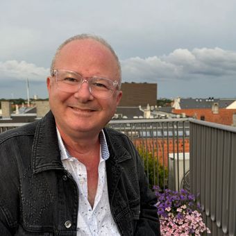 A man wearing glasses and a black jacket is sitting on a balcony.