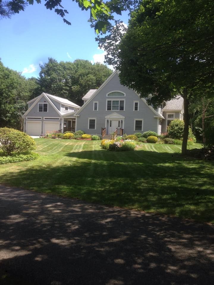 Gray house with two-car garage, trimmed lawn, and trees, viewed from a shaded driveway on a sunny day.