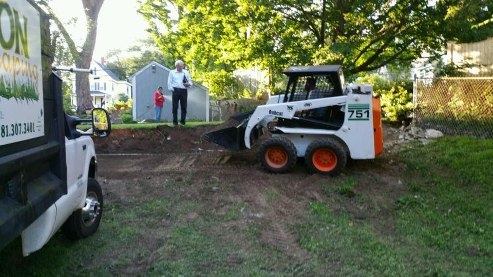 Bobcat skid steer scoops dirt in a yard next to a landscaping truck, two people watch.