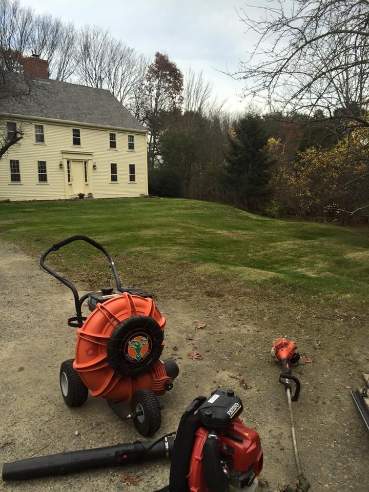 Orange leaf blower and trimmer on a gravel driveway, large yellow house in background, green lawn.