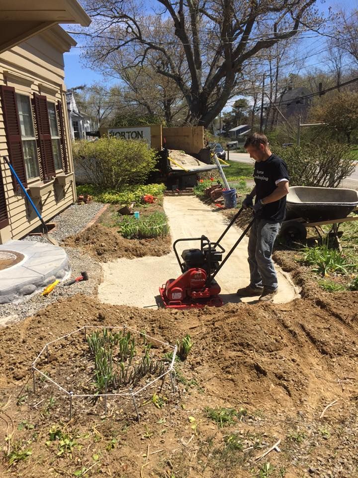 Man using a vibratory plate compactor to compact soil for a walkway next to a house.