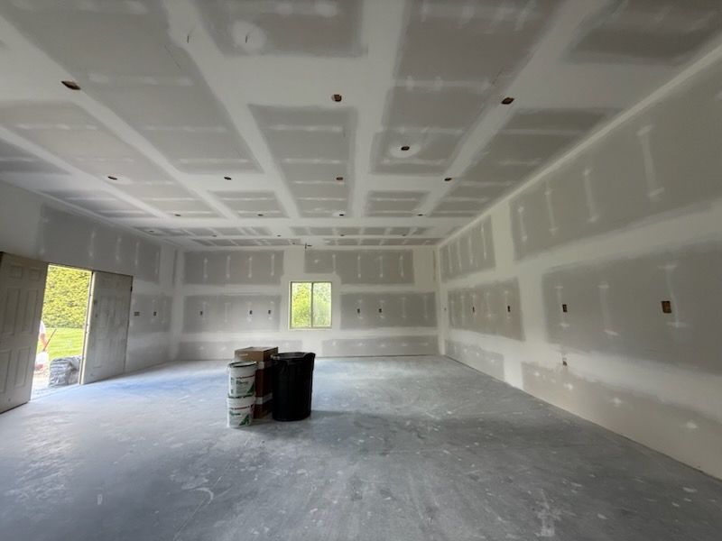 Interior of a room under construction, with drywall on walls and ceiling, a window, and barrels in the center.
