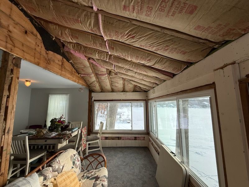 Sunroom interior with exposed ceiling insulation and windows overlooking snow-covered scenery. Dining room visible.
