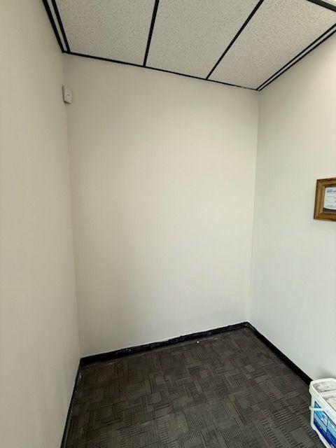 A small, empty office corner with off-white walls, dark gray carpet, a drop ceiling, and a framed picture on the wall.