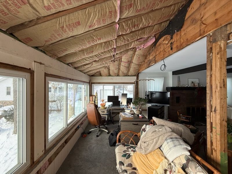 Interior view of a sunroom with exposed insulation, windows, desk, and furniture. Snowy outdoor view.