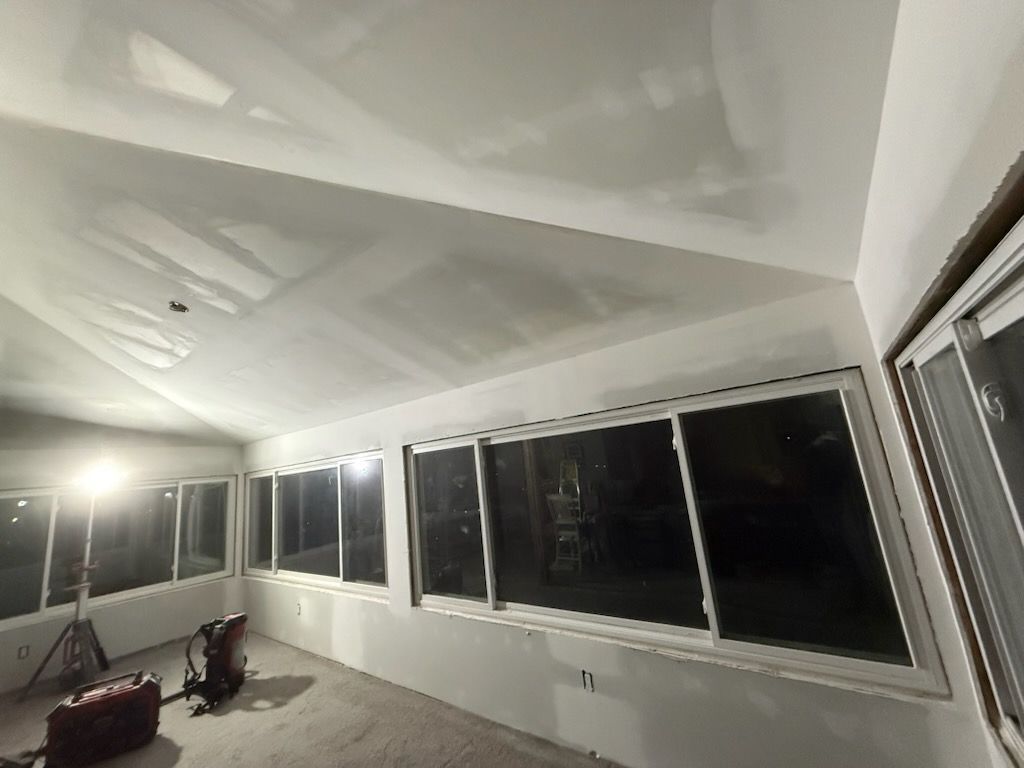 Interior view of a room with unfinished drywall and large windows, lit by a work light.