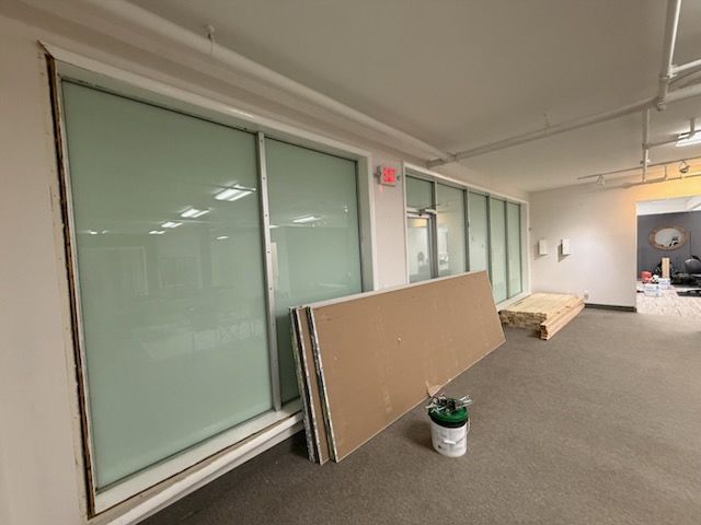 A large office space under construction with frosted glass partitions, stacks of drywall, and a paint bucket on the floor.