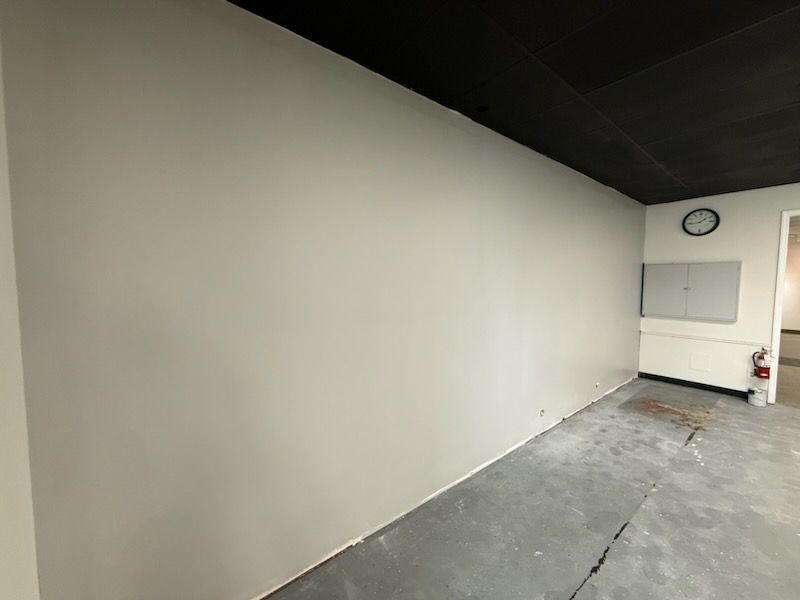 An interior view of a room with a large, light grey wall, a black ceiling, and unfinished concrete flooring.