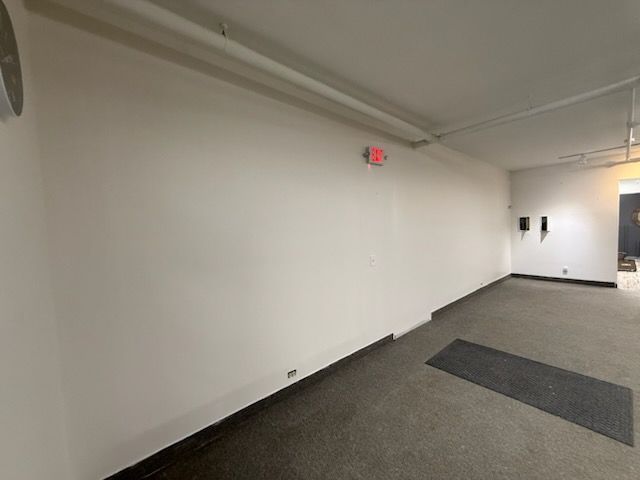 An expansive, empty white room with a gray carpeted floor, a long horizontal pipe on the ceiling, and an exit sign.