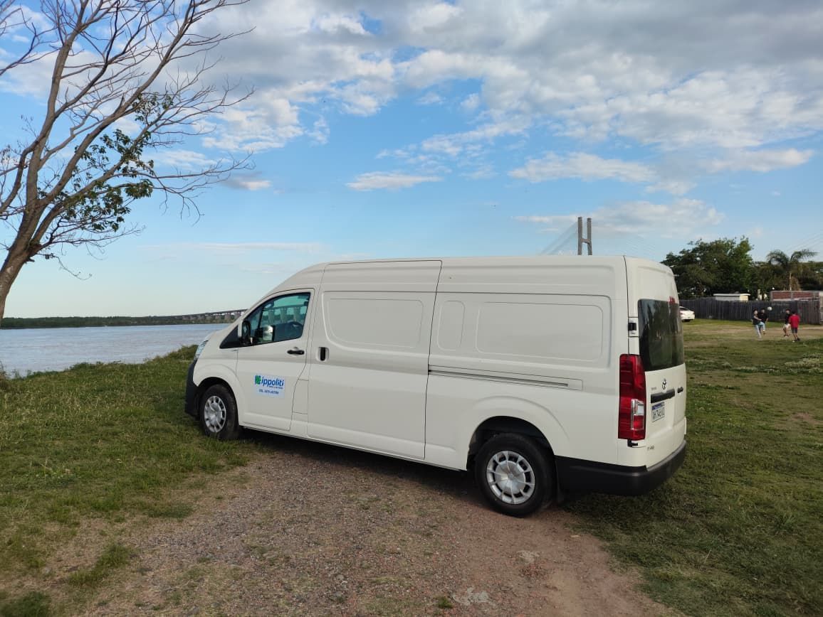 Furgoneta blanca estacionada en la orilla de hierba junto al agua, bajo un cielo azul nublado.