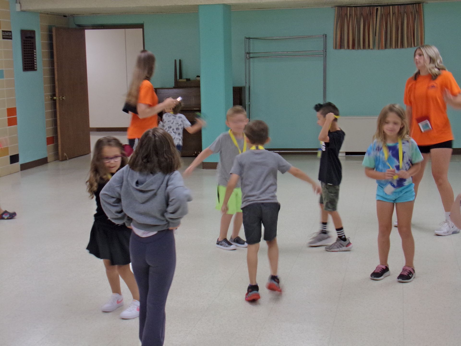 Children and counselors in a room, some wearing medals, gesturing, laughing. Bright room, teal walls, activity.