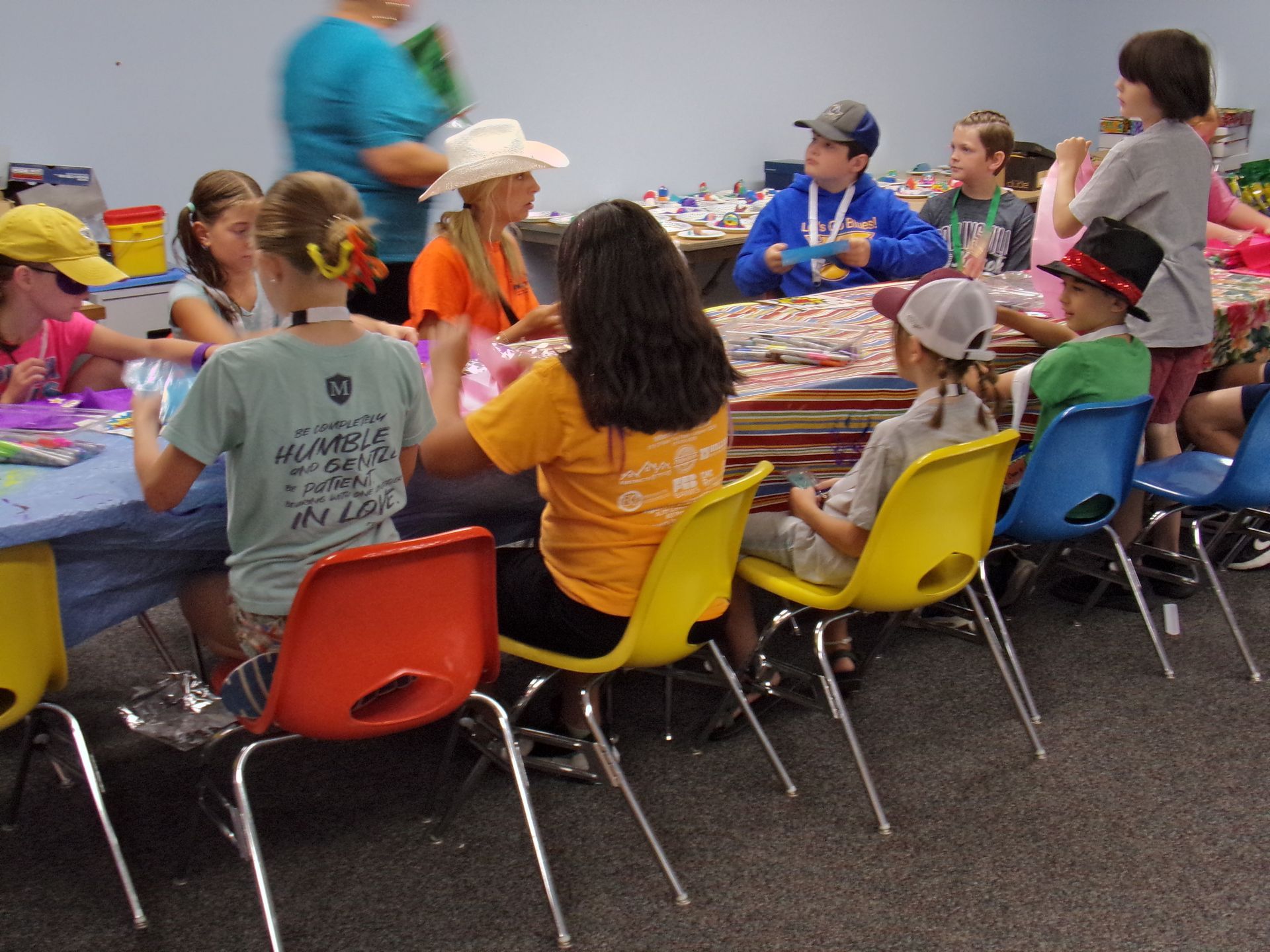 Children and adults sit around tables crafting. Various colorful chairs. Workshop setting.