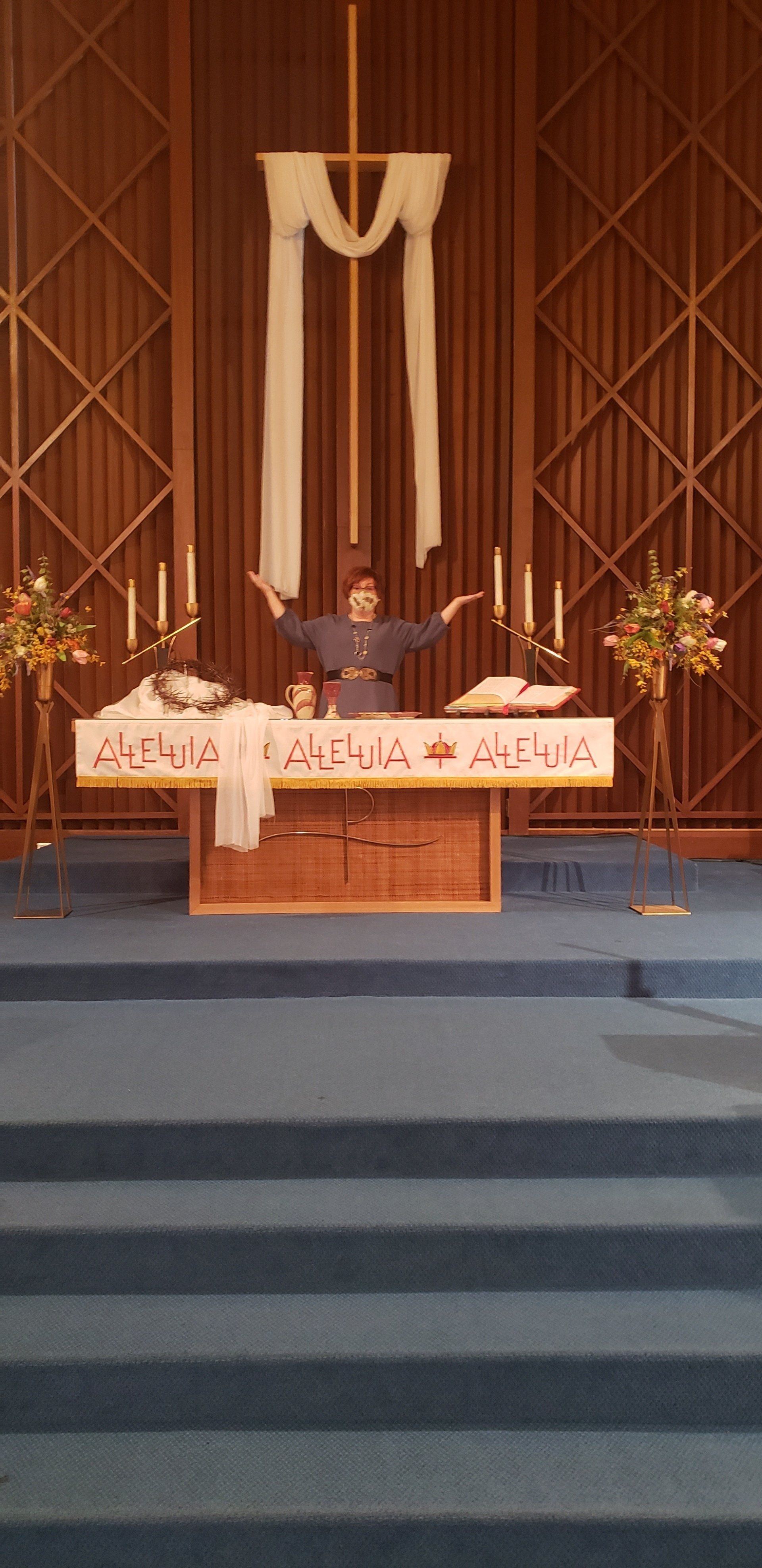 A person with arms raised in a church, standing behind a decorated altar with cross.