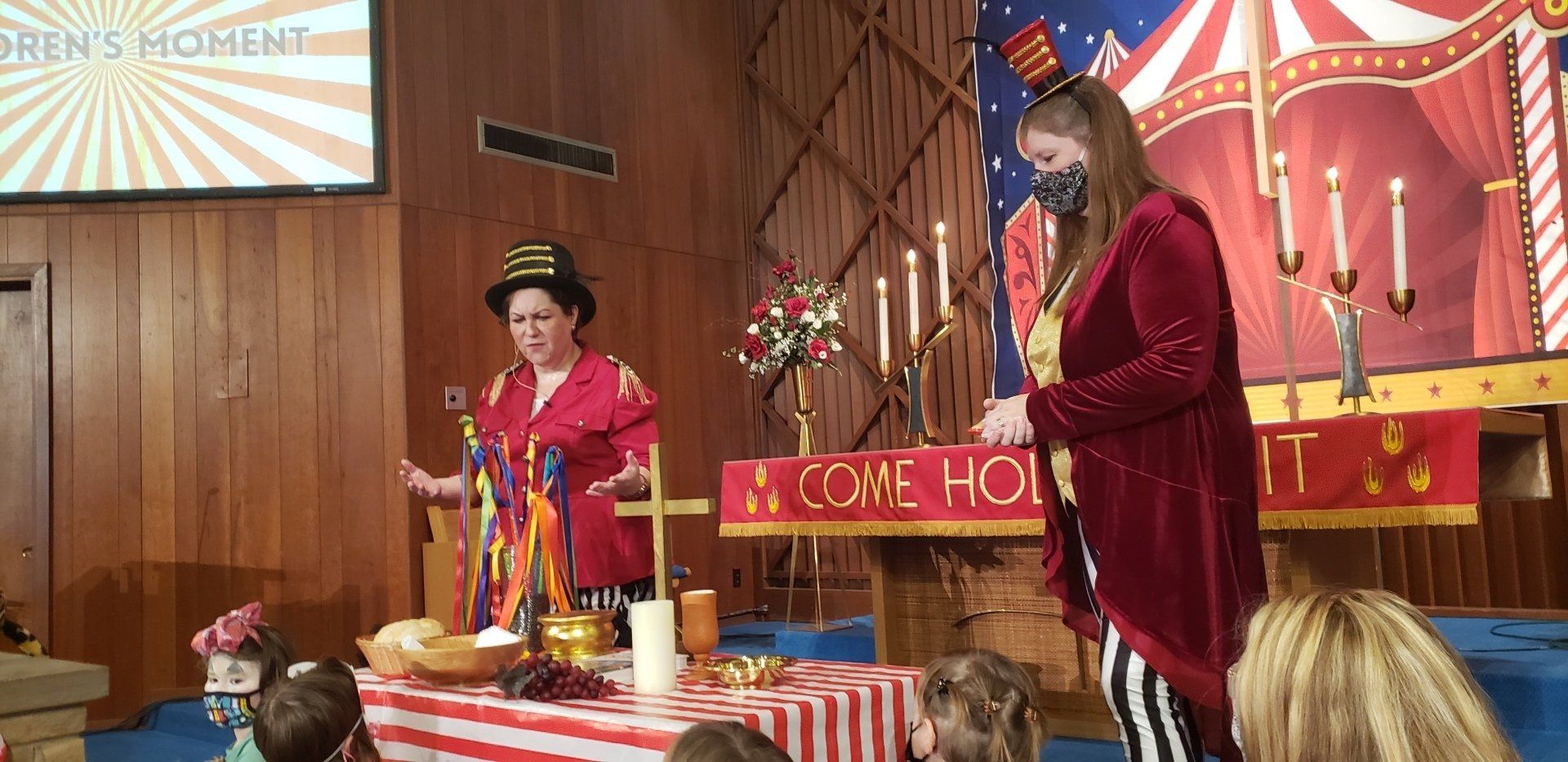 Two women in circus costumes at a church altar. One is speaking, the other standing with a mask.