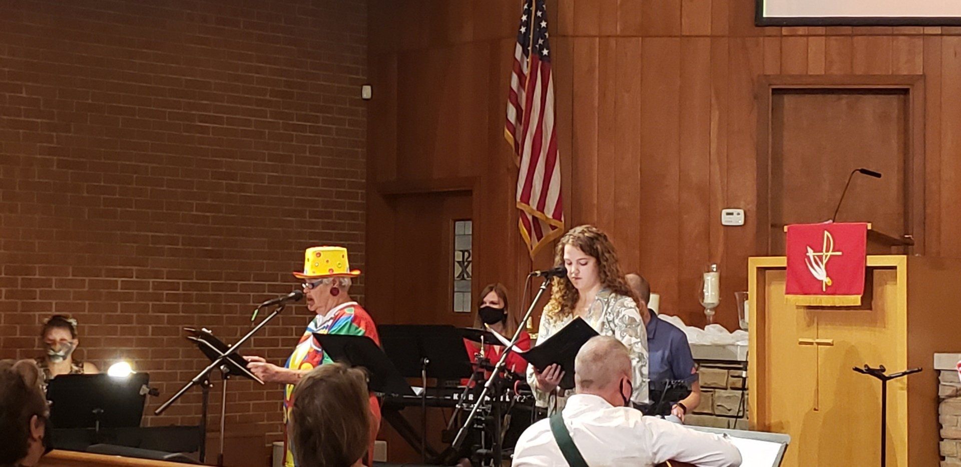 A band performs on a stage in a church. The singer in a yellow hat plays guitar. Others sing, holding music.