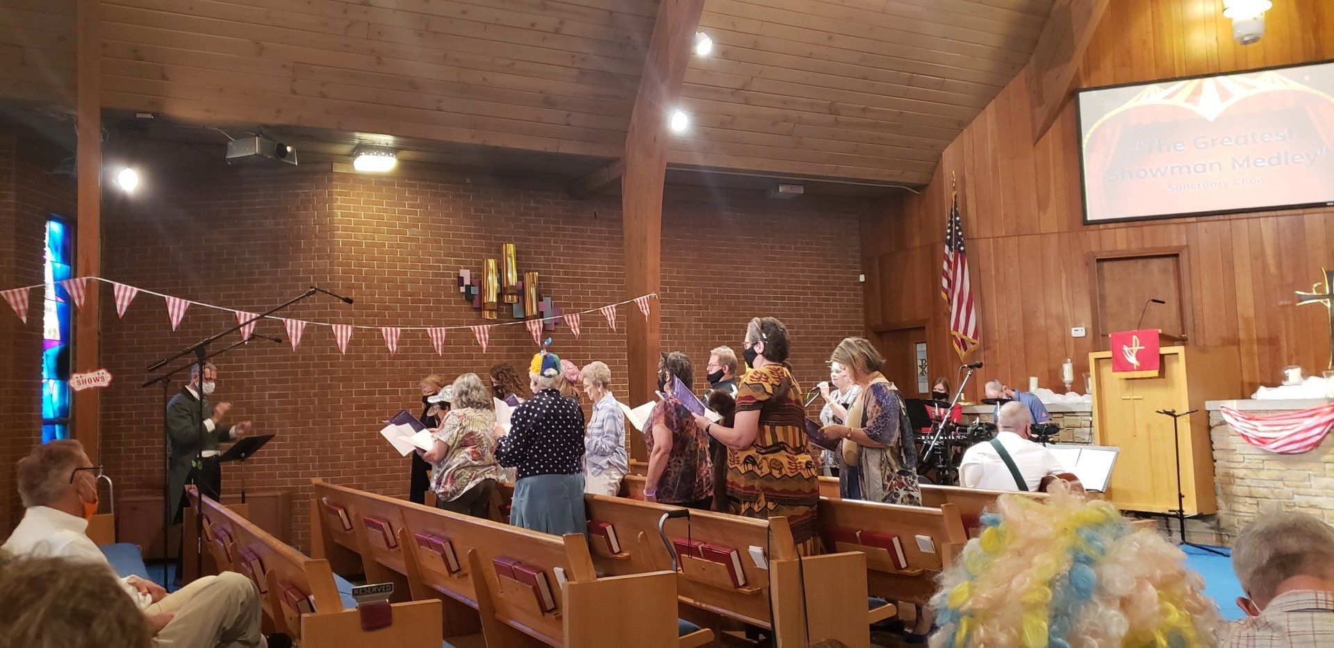 People singing in a church. Some are wearing masks. Banners and a US flag are visible.