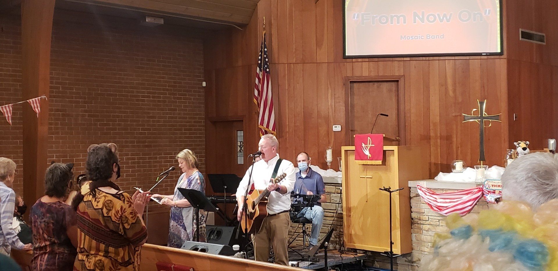 A worship band performs in a church, with a guitarist singing. Audience clapping. American flag hangs.