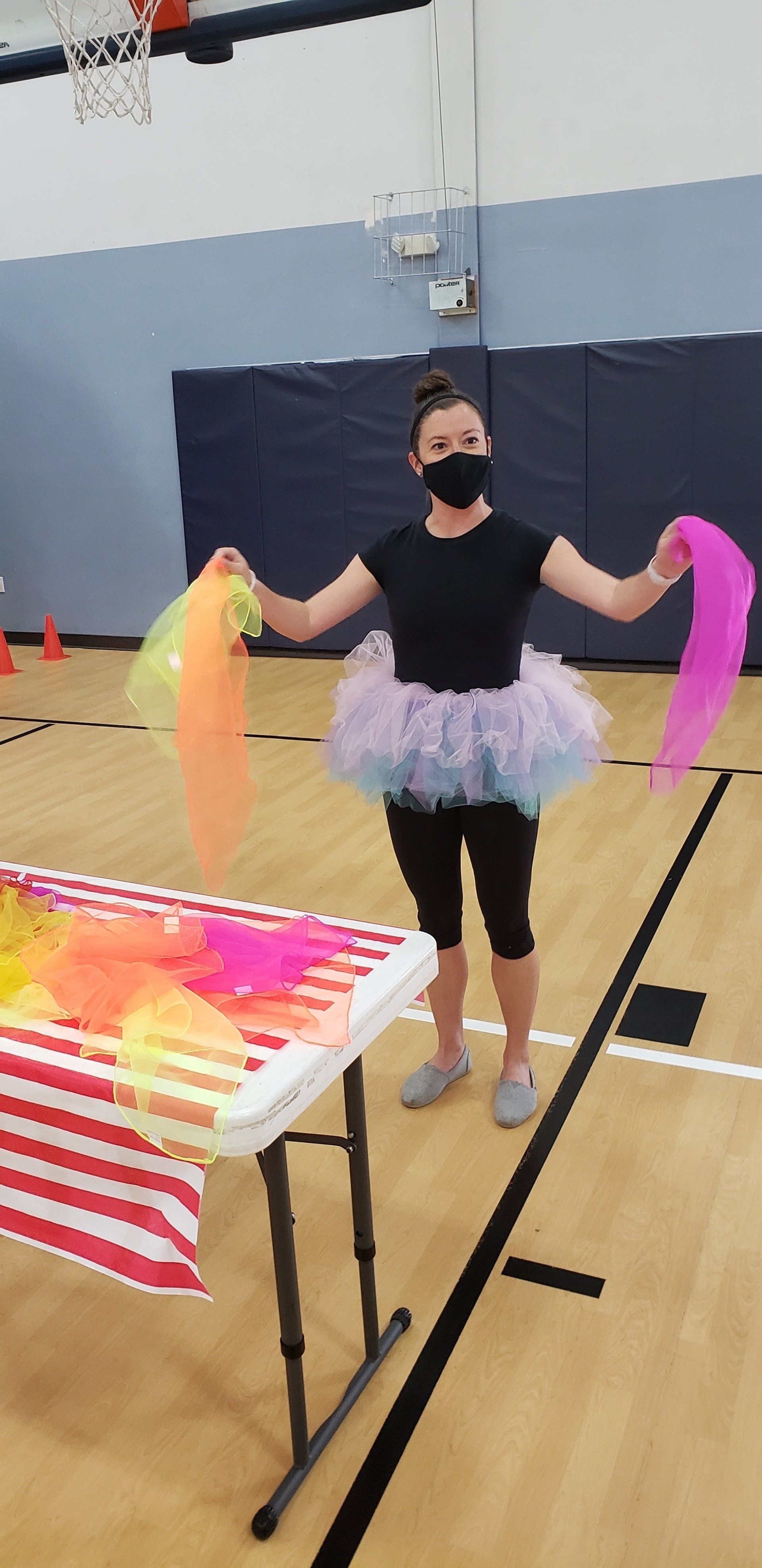 A woman wearing a tutu and mask holds colorful scarves in a gym.