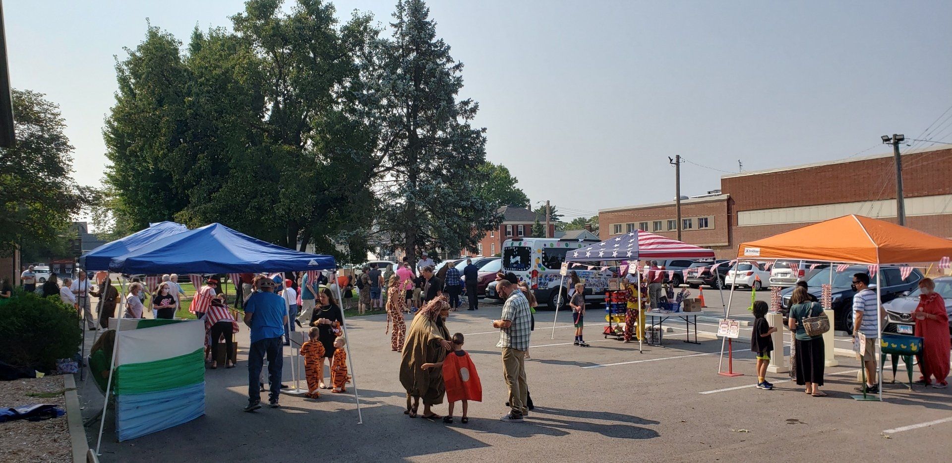 People gathered outdoors under tents, likely at an event in a parking lot, with a brick building in the background.