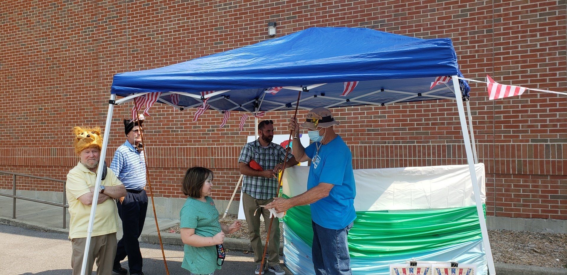 People under a blue canopy. A man in a blue shirt hands something to a child. Others stand nearby.